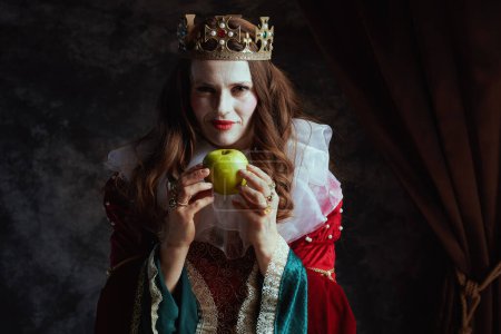 Photo for Insidious medieval queen in red dress with green apple, white collar and crown on dark gray background. - Royalty Free Image