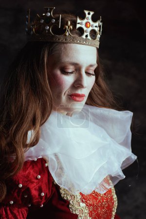 Photo for Pensive medieval queen in red dress with white collar and crown on dark gray background. - Royalty Free Image