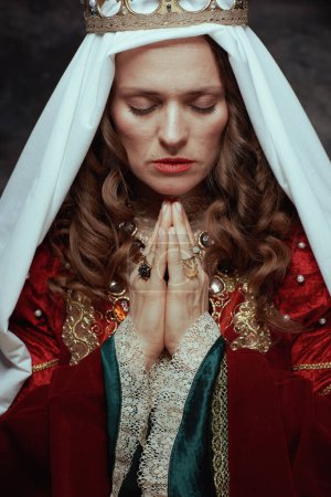 Téléchargez les photos : Gros plan sur la reine médiévale en robe rouge avec voile et couronne priant sur fond gris foncé. - en image libre de droit