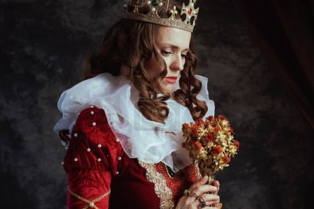 Foto de Reina medieval en vestido rojo con flor seca, cuello blanco y corona sobre fondo gris oscuro. - Imagen libre de derechos