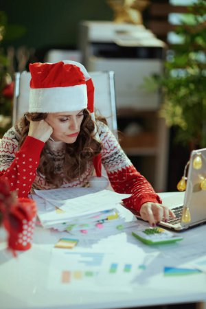 Photo for Christmas time. stressed stylish small business owner woman in santa hat and red Christmas sweater with documents and laptop working in modern green office with Christmas tree. - Royalty Free Image