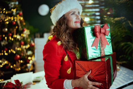 Téléchargez les photos : Noël. souriant élégant 40 ans propriétaire de petite entreprise femme en santa chapeau et veste rouge avec des boîtes cadeaux dans le bureau vert moderne avec arbre de Noël. - en image libre de droit