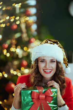Téléchargez les photos : Noël. souriant moderne propriétaire de petite entreprise femme en santa chapeau et veste rouge avec boîte cadeau dans le bureau vert moderne avec arbre de Noël. - en image libre de droit