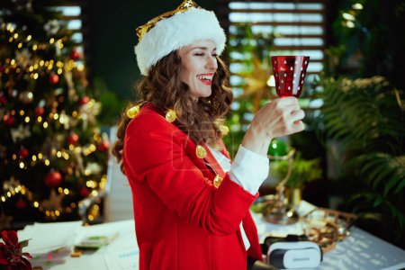 Foto de En Navidad. feliz mujer de negocios elegante de mediana edad en sombrero de santa y chaqueta roja con copa de vino en la oficina verde moderna con árbol de Navidad. - Imagen libre de derechos
