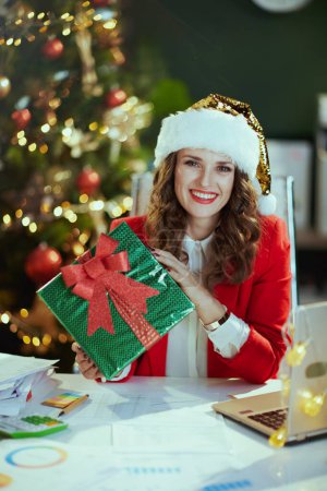 Téléchargez les photos : Noël. heureuse élégante propriétaire de petite entreprise d'âge moyen femme en santa chapeau et veste rouge avec boîte cadeau dans le bureau vert moderne avec arbre de Noël. - en image libre de droit