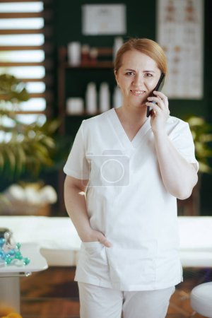 Photo for Healthcare time. happy female massage therapist in massage cabinet speaking on a smartphone. - Royalty Free Image