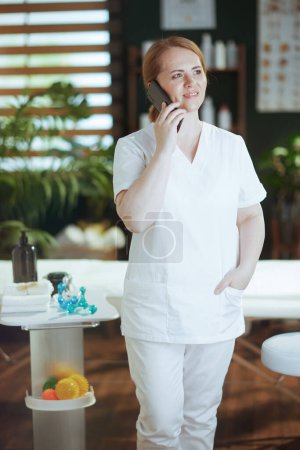 Foto de Tiempo de atención médica. sonriente masaje terapeuta mujer en spa salón hablando en un teléfono inteligente. - Imagen libre de derechos