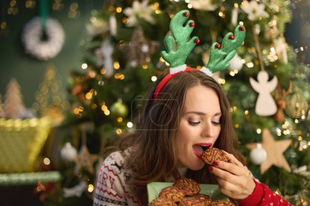 Téléchargez les photos : Noël. élégante femme d'âge moyen avec biscuit de Noël et drôle de cerceau de bois de renne en pull de Noël traditionnel près de l'arbre de Noël à la maison moderne. - en image libre de droit