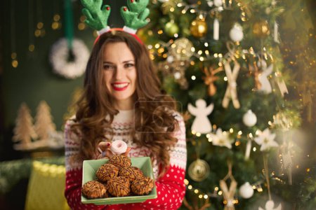 Photo for Christmas time. happy elegant 40 years old housewife with Christmas cookie and funny reindeer antlers headpiece hoop in traditional Christmas sweater near Christmas tree in the modern living room. - Royalty Free Image