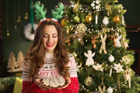 Photo for Christmas time. happy trendy housewife with Christmas cookie and funny reindeer antlers headpiece hoop in traditional Christmas sweater near Christmas tree in the modern house. - Royalty Free Image