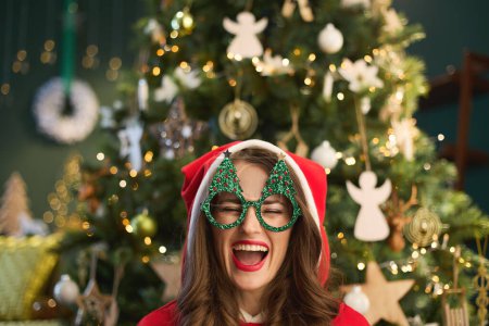Foto de En Navidad. alegre mujer de moda con sombrero de Santa y gafas de árbol de Navidad divertido cerca del árbol de Navidad en la sala de estar moderna. - Imagen libre de derechos