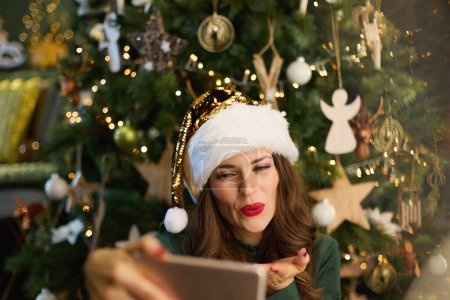 Foto de En Navidad. feliz mujer elegante con sombrero de Santa en vestido verde que tiene video chat en un teléfono inteligente cerca del árbol de Navidad en el hogar moderno. - Imagen libre de derechos