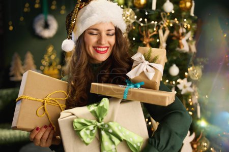 Foto de En Navidad. sonriente elegante ama de casa con sombrero de Santa y eco empaquetado cajas de regalo en vestido verde cerca del árbol de Navidad en el hogar moderno. - Imagen libre de derechos