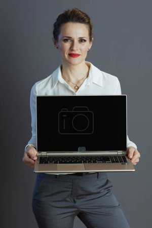 Téléchargez les photos : Femme d'affaires moderne de 40 ans en chemisier blanc montrant écran blanc d'ordinateur portable isolé sur fond gris. - en image libre de droit
