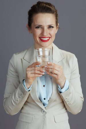 Téléchargez les photos : Heureuse élégante propriétaire de petite entreprise femme dans un costume d'affaires léger avec verre d'eau isolé sur gris. - en image libre de droit