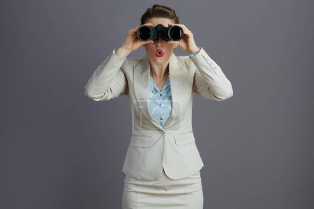 Foto de Sorprendida empleada femenina elegante en un traje de negocios ligero con prismáticos aislados en gris. - Imagen libre de derechos