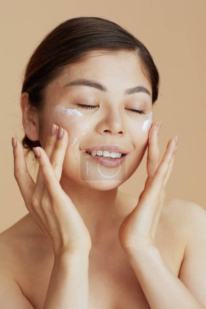 Téléchargez les photos : Portrait de femme asiatique moderne avec crème faciale sur le visage sur fond beige. - en image libre de droit