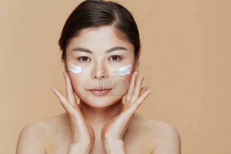 Foto de Mujer asiática moderna con crema facial en la cara aislada sobre fondo beige. - Imagen libre de derechos