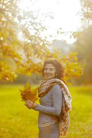 Foto de Hola otoño. Retrato de la feliz y elegante mujer de 40 años en el parque con hojas de otoño y bufanda. - Imagen libre de derechos
