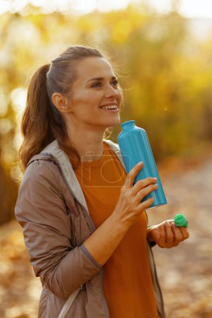 Foto de Hola otoño. sonriente mujer moderna en ropa de fitness en el parque con botella de agua. - Imagen libre de derechos