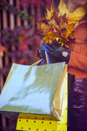 Photo for Hello november. Closeup on elegant female in brown trench coat with shopping bags and autumn yellow leaves in the city. - Royalty Free Image