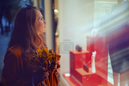 Foto de Hola septiembre. elegante mujer de mediana edad en gabardina naranja con hojas amarillas de otoño cerca de la tienda en la ciudad. - Imagen libre de derechos