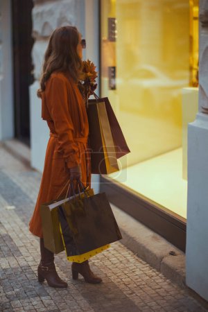 Foto de Hola octubre. Vista lateral de la joven hembra en gabardina naranja con bolsas de compras y hojas amarillas de otoño cerca de la tienda en la ciudad. - Imagen libre de derechos