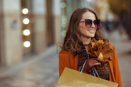 Téléchargez les photos : Bonjour, novembre. heureuse jeune femme en trench-coat orange avec des sacs à provisions et des feuilles jaune automne dans la ville. - en image libre de droit