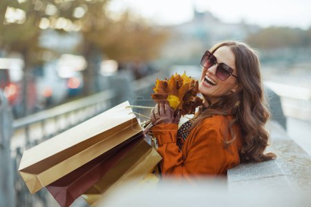 Foto de Hola octubre. joven hembra sonriente en gabardina marrón con bolsas de compras y hojas amarillas otoñales en la ciudad. - Imagen libre de derechos