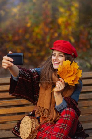 Foto de Hola otoño. sonriente joven hembra en sombrero rojo con hojas de otoño, bufanda y bolso teniendo video reunión mientras está sentado en el banco en el parque de la ciudad. - Imagen libre de derechos