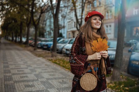 Foto de Hola otoño. sonriente mujer de 40 años en sombrero rojo con hojas de otoño y bufanda caminando por la ciudad. - Imagen libre de derechos