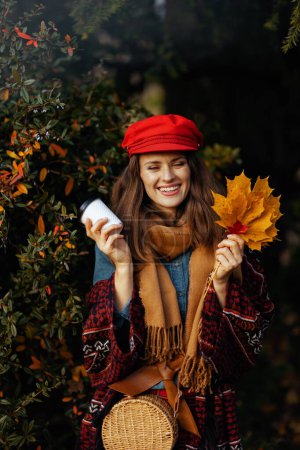 Foto de Hola otoño. mujer moderna feliz en sombrero rojo con hojas de otoño, bufanda, bolsa y café en el parque de la ciudad. - Imagen libre de derechos