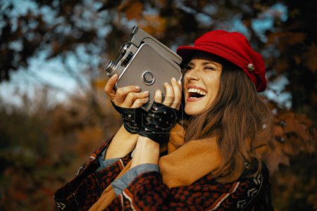 Téléchargez les photos : Bonjour l'automne. femme élégante et souriante en chapeau rouge avec caméra vidéo rétro, écharpe et gants dans le parc de la ville. - en image libre de droit
