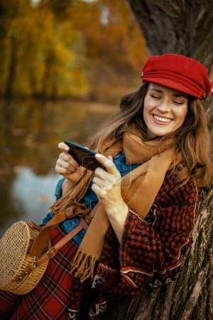Photo for Hello autumn. smiling modern female in red hat with scarf and bag using smartphone near tree in the city park. - Royalty Free Image