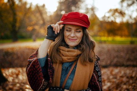 Hallo Herbst. Lächelnde moderne 40-jährige Frau mit roter Mütze, Schal und Handschuhen im Stadtpark.