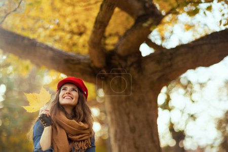 Foto de Hola otoño. mujer de mediana edad feliz de moda en jeans camisa y sombrero rojo con hoja de otoño, bufanda y guantes cerca del árbol en el parque de la ciudad. - Imagen libre de derechos