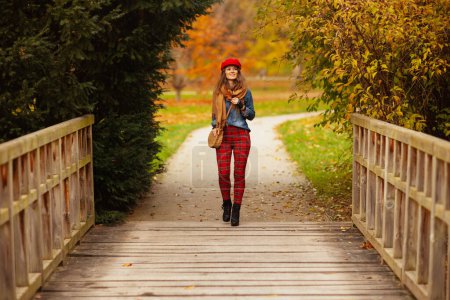 Téléchargez les photos : Bonjour l'automne. Portrait pleine longueur de femme souriante à la mode en jeans chemise et chapeau rouge avec feuille d'automne, écharpe et sac à pied dans le parc de la ville. - en image libre de droit