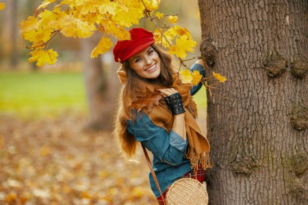 Foto de Hola otoño. Retrato de mujer con estilo feliz en camisa vaquera y sombrero rojo con hojas de otoño, bufanda, guantes y bolsa cerca del árbol en el parque de la ciudad. - Imagen libre de derechos