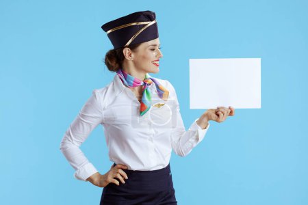 Téléchargez les photos : Femme hôtesse de l'air moderne souriante sur fond bleu en uniforme montrant feuille de papier vierge a4. - en image libre de droit