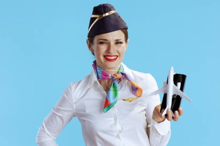 Photo for Smiling elegant stewardess woman isolated on blue background in uniform showing a little airplane and smartphone. - Royalty Free Image