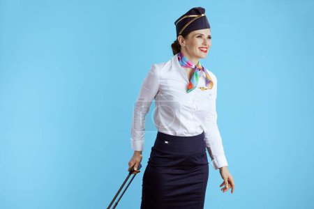 Foto de Sonriente elegante azafata mujer sobre fondo azul en uniforme con bolsa de ruedas mirando a la distancia. - Imagen libre de derechos