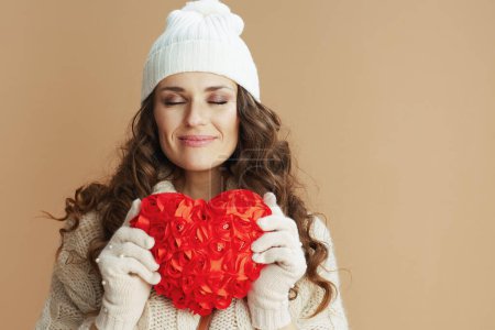 Téléchargez les photos : Bonjour l'hiver. femme détendue et tendance en pull beige, mitaines et chapeau isolés sur fond beige avec coeur rouge. - en image libre de droit