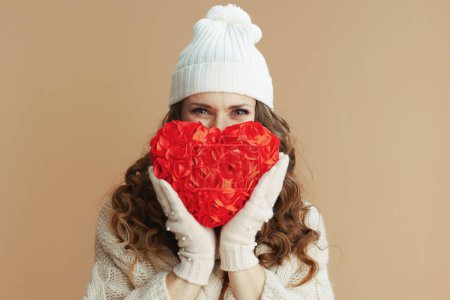 Téléchargez les photos : Bonjour l'hiver. élégante femme en pull beige, mitaines et chapeau sur fond beige avec coeur rouge. - en image libre de droit