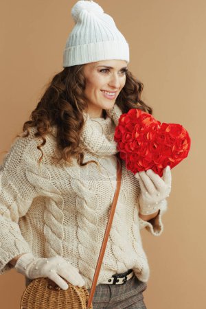 Téléchargez les photos : Bonjour l'hiver. femme souriante élégante de 40 ans en pull beige, mitaines et chapeau avec coeur rouge et sac à main isolé sur beige. - en image libre de droit