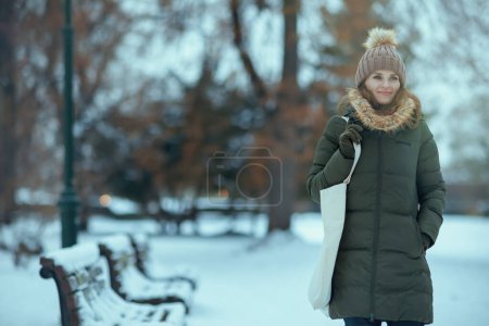 Foto de Feliz mujer moderna en abrigo verde y sombrero marrón al aire libre en el parque de la ciudad en invierno con manoplas y gorro sombrero. - Imagen libre de derechos