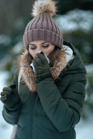 Photo for Unhappy modern middle aged woman in green coat and brown hat outdoors in the city park in winter with mittens, napkin blowing nose and beanie hat. - Royalty Free Image