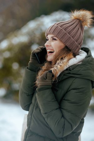 Foto de Mujer moderna feliz en abrigo verde y sombrero marrón al aire libre en el parque de la ciudad en invierno con manoplas y gorro sombrero hablando en un teléfono inteligente. - Imagen libre de derechos