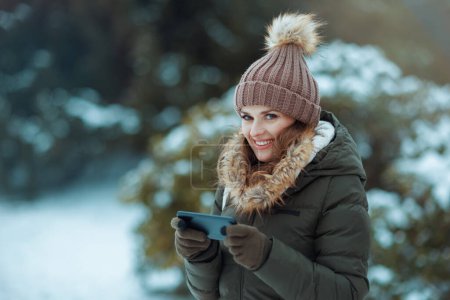 Foto de Sonriente hembra moderna en abrigo verde y sombrero marrón al aire libre en el parque de la ciudad en invierno con manoplas y gorro enviando un mensaje de texto utilizando un teléfono inteligente cerca de las ramas nevadas. - Imagen libre de derechos
