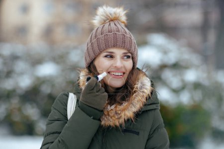 Photo for Happy modern middle aged woman in green coat and brown hat outdoors in the city park in winter with mittens, hygienic lipstick and beanie hat. - Royalty Free Image