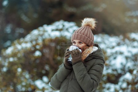 Téléchargez les photos : Coûteuse femelle moderne en manteau vert et chapeau brun à l'extérieur dans le parc de la ville en hiver avec des mitaines, une tasse de cacao chaud et un bonnet près des branches enneigées. - en image libre de droit